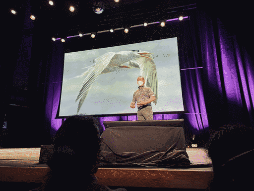 Ed Yong talking about birding, standing in front of a beautiful photo he took of a bird skimming across the water.